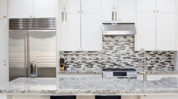 A brightly lit kitchen with granite countertops, white cabinets, stainless steel appliances, and a tiled backsplash.
