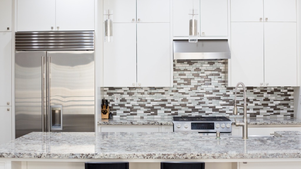 A brightly lit kitchen with granite countertops, white cabinets, stainless steel appliances, and a tiled backsplash.