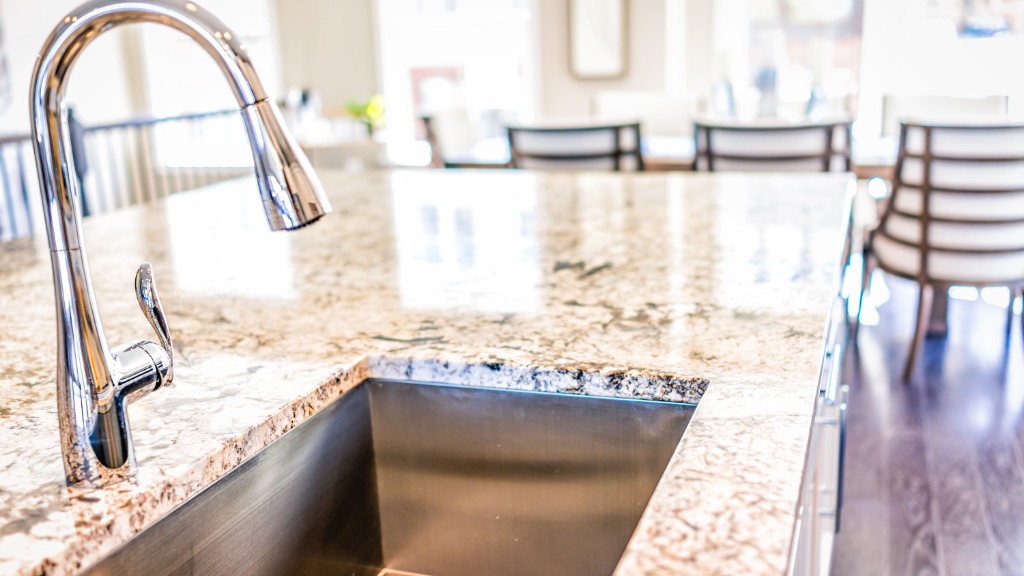 A close-up of a deep stainless steel sink in a kitchen. Bright granite countertops surround the sink basin.